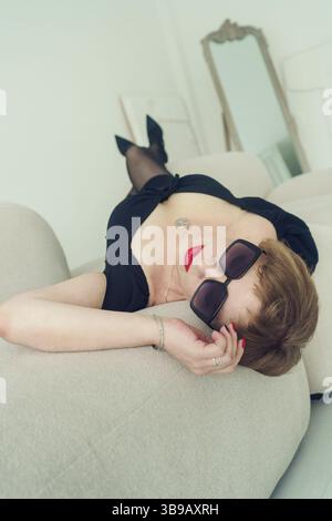 una donna bruna adulta con i capelli corti in un abito nero è sdraiata sul divano appoggiata alla schiena Foto Stock