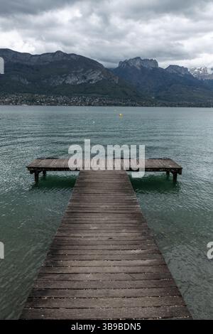 Un molo di legno si estende nelle acque del lago di Annecy in Francia, con viste spettacolari delle montagne alpine sullo sfondo e nuvole scure sopra Foto Stock