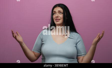 La donna esprime frustrazione su sfondo rosa isolato, indossa una camicia azzurra, taglia, capelli neri lunghi e curvati, stile casual con una borsa chiusa Foto Stock