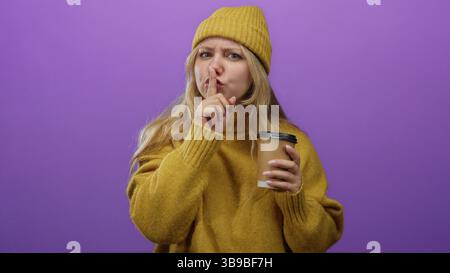 Una donna in giallo sembra curiosa con un dito sulle labbra che tiene il caffè contro una vivace parete viola. Foto Stock