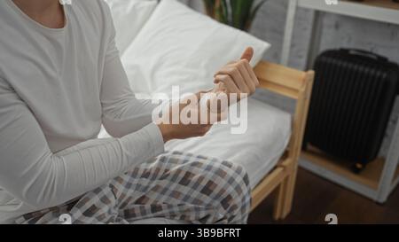 Giovane uomo seduto sul letto in camera da letto con pigiama, polso a controllo, con valigia sullo sfondo, che mostra un'atmosfera rilassata al mattino all'interno. Foto Stock