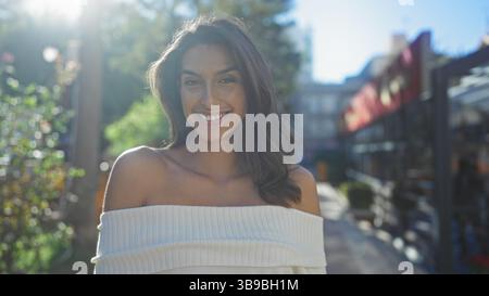 Donna in piedi nel parco urbano sorridente sotto la luce del sole con alberi ed edifici sullo sfondo che indossano la parte superiore bianca trasuda bellezza e fiducia in una città st Foto Stock