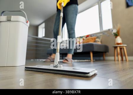 Donna che pulisce, pulisce eleganti pavimenti in legno duro in un ambiente moderno. Ragazza che indossa guanti gialli. La casalinga utilizza prodotti di pulizia professionali: Mocio, secchio di plastica con strizzatore in un accogliente salotto Foto Stock