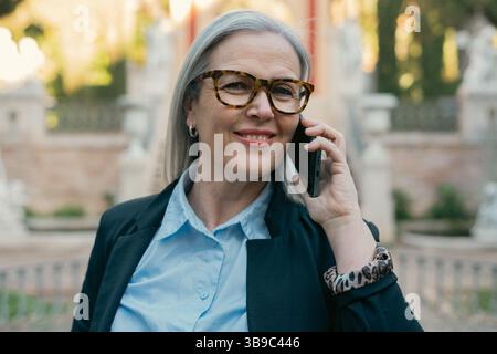Una donna matura e sicura di sé impegnata in una conversazione telefonica mentre si trova in un ambiente urbano Foto Stock