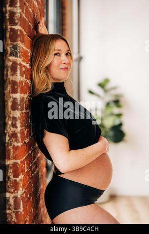 Donna incinta in nero appoggiata contro le pareti di mattoni in casa Foto Stock