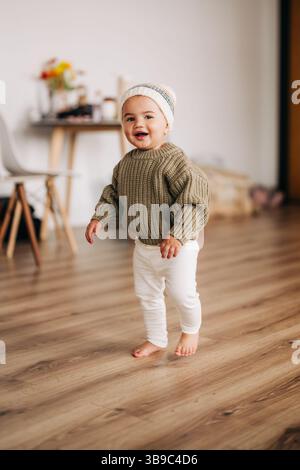 Bimbo sorridente in maglia e berretto che cammina in casa Foto Stock