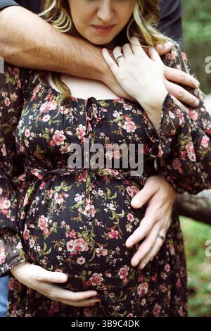 Donna incinta alla moda abbracciata da un forte marito in un abito floreale Foto Stock