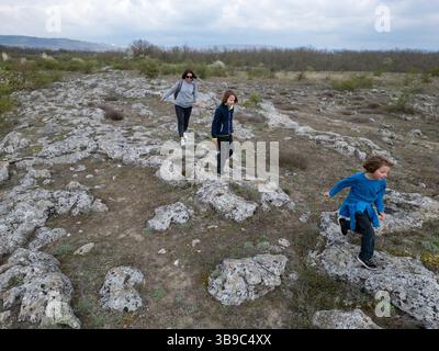 Madre e due bambini esplorano il terreno roccioso in un paesaggio arido e semi-desertico. I bambini giocano su formazioni di pietra mentre la madre guarda, enjo Foto Stock