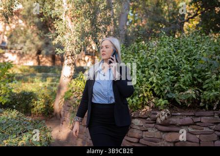 Donna d'affari che cammina in un parco e parla con lo smartphone Foto Stock