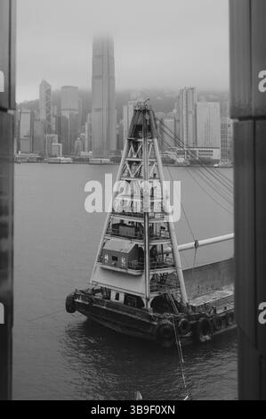 Vista monocromatica di una chiatta costruita incorniciata contro lo skyline avvolto dalla nebbia dell'Isola di Hong Kong Foto Stock