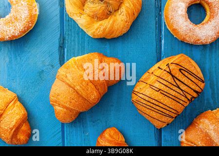 Croissant appena sfornati, ciambelle, pain au chocolat e altri dolci dolci sono disposti su un vivace tavolo di legno blu, creando un'allettante colazione Foto Stock