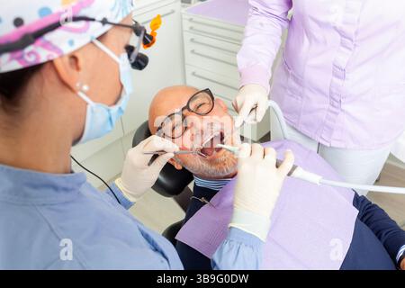 Dentista che indossa lenti d'ingrandimento che esegue un controllo dentale su un paziente di sesso maschile senior con l'assistenza di un'infermiera dentale Foto Stock