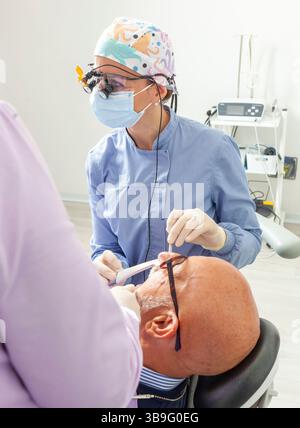 Dentista donna che indossa lenti d'ingrandimento che esegue un controllo dentale su un anziano sdraiato su una sedia dentista Foto Stock