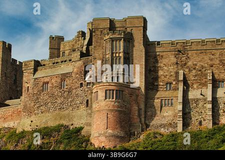 Un grande e imponente castello in pietra con torri, mura e finestre multiple, visto da un'angolazione bassa. Vegetazione verde alla base del castello di Bamburgh, Regno Unito. Foto Stock