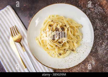 Un piatto di tagliatelle guarnite con tartufo nero, elegantemente collocato su un tavolo rustico. Spara con utensili dorati e un tovagliolo a strisce per dini Foto Stock