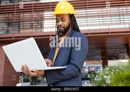 Uomo d'affari cubano che indossa un elmetto giallo e una tuta marina, impegnato con un computer portatile fuori da un edificio moderno. Rappresenta l'integrazione tecnologica in const Foto Stock
