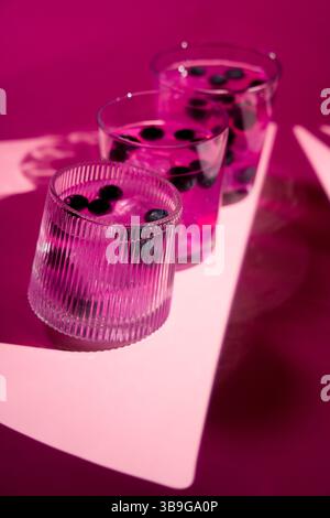 Tre bicchieri trasparenti pieni di una vivace bevanda rosa e mirtilli galleggianti si trovano su una superficie rosa impressionante, che getta ombre artistiche. I composi Foto Stock