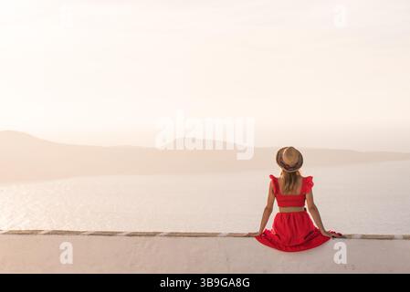 Una donna con un abito rosso siede su una parete bianca, di fronte al tranquillo paesaggio marino di Plaka, isola di Milos, Grecia. Indossa un cappello, catturando un momento di pace Foto Stock