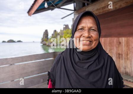 Una donna allegra in un hijab nero sorride brillantemente, in piedi su un ponte di legno da splendide formazioni d'acqua e di roccia dell'Indonesia, sotto un tetto rustico Foto Stock