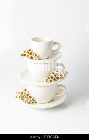 Tazze e piattini bianchi impilati adornati con delicati fiori primaverili. Un'estetica pulita e minimalista in un ambiente luminoso migliora il fascino visivo di Foto Stock