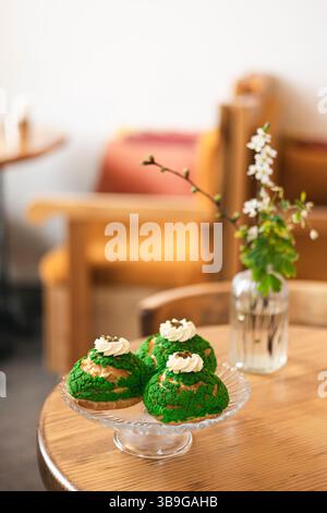 Tre dolci matcha choux su un piatto di vetro mettono in risalto questo intimo ambiente da caffetteria con un tavolo in legno e un semplice decor floreale, creando un invitante e chicco Foto Stock