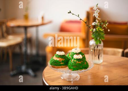 Un piatto di vetro con dolci matcha choux con panna montata siede su un tavolo di legno in un intimo caffè. Sullo sfondo, mobili sfocati e un fiore V Foto Stock
