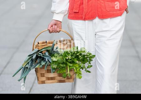 Una donna alla moda con giacca rossa e pantaloni bianchi porta un cesto in tessuto pieno di porri e prezzemolo all'aperto, enfatizzando i prodotti freschi e la moda Foto Stock