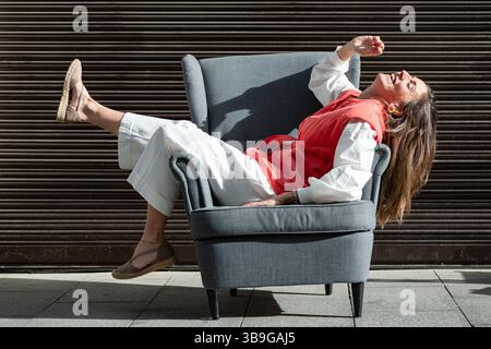Una donna alla moda con pantaloni bianchi e un gilet rosso ride allegramente mentre si sdraia su una poltrona grigia. È seduta all'aperto con uno sfondo scuro. Foto Stock