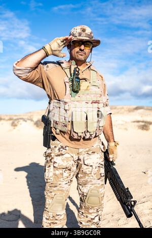 Un soldato in piena mimetizzazione nel deserto, dotato di equipaggiamento tattico e fucile, saluta con sicurezza contro un cielo azzurro. La scena si trasmette Foto Stock