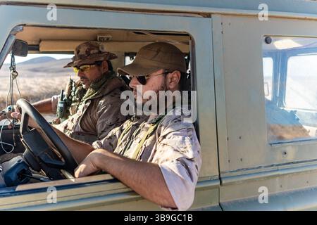 Due soldati militari con equipaggiamento tattico siedono all'interno di un veicolo corazzato che guida attraverso un terreno desertico, concentrandosi sulla loro missione. Il paesaggio asciutto Foto Stock
