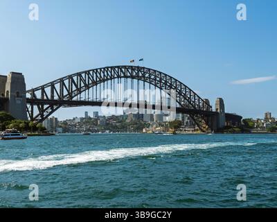 Harbour Bridge si estende attraverso una vivace città, con barche e skyline della città sullo sfondo, sotto un cielo blu brillante a Sidney. Foto Stock