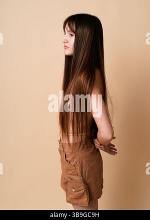 Vista laterale di una giovane donna bruna con i capelli lunghi che guarda lontano in piedi sullo sfondo di uno studio beige, indossando un elegante abito marrone Foto Stock