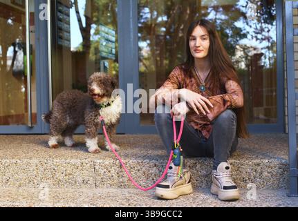 Una giovane donna siede sui gradini accanto a un cane con un guinzaglio rosa, godendosi una giornata di sole. La scena cattura un momento di compagnia e relax all'aperto Foto Stock