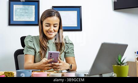 Una nutrizionista si siede alla sua scrivania, sorridendo mentre utilizza uno smartphone, con un laptop aperto. I certificati sul muro indicano la sua qualifica professionale Foto Stock