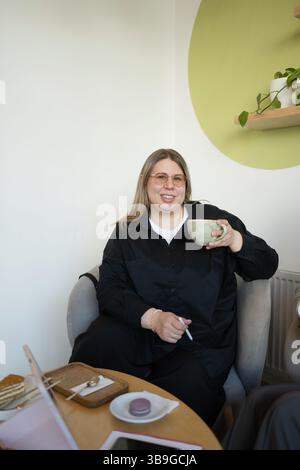 Una donna con un vestito nero si rilassa con una tazza di caffè in un caffè luminoso ed elegante. Si siede comodamente, circondata da dessert sul tavolo, creando Foto Stock
