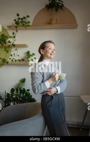 Una donna si trova in un elegante caffè, sorridendo e tenendo in mano una tazza di caffè. Gli interni luminosi e moderni presentano piante e scaffali in legno, creando un rela Foto Stock