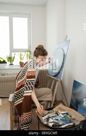 Un artista in uno studio accogliente e pieno di luce lavora su una tela con pennelli e tubi nelle vicinanze, creando un'atmosfera serena e concentrata. Foto Stock