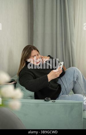 Una donna allegra siede comodamente su un divano color pastello, utilizzando il suo smartphone e sorridendo. Vestita in modo informale con un maglione nero e un paio di jeans, le piace Foto Stock