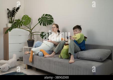 Due bambini siedono su un divano grigio, giocando ai videogiochi con i controller in mano. I peluche riposano sulle loro laccature e le piante verdi decorano la stanza, cre Foto Stock