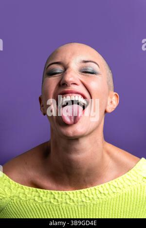 Una donna di mezza età con la testa rasata si attacca con gioia sulla lingua, trasmettendo un senso di libertà e audacia. Il suo maglione verde brillante contrasta lo spirito Foto Stock