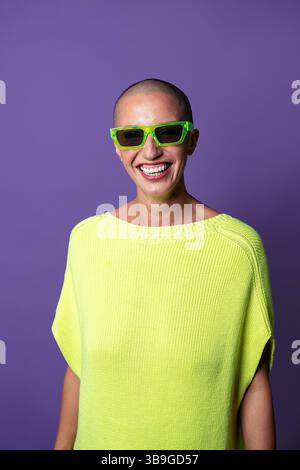 Una donna di mezza età con testa rasata, occhiali da sole verdi al neon e un maglione giallo brillante, sorride con energia vibrante contro una solida b viola Foto Stock