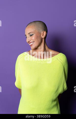 Una donna di mezza età con la testa rasata sorride con fiducia su un vibrante sfondo viola, indossando un maglione giallo brillante che simboleggia gli uomini potenti Foto Stock