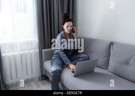 Una donna in pigiama e una maschera facciale si siede su un divano grigio, multitasking tra un notebook e un telefono. La sala dispone di luce naturale soffusa Foto Stock