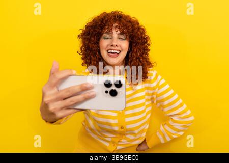 Una donna sorridente con i capelli ricci rossi in una camicia a righe gialle scatta un selfie su uno sfondo giallo brillante. La sua espressione allegra irradia gioia Foto Stock