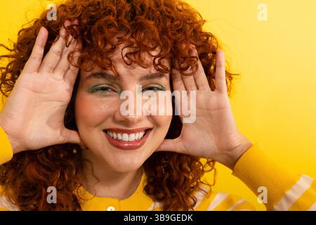 Una donna sorridente con i capelli rossi ricci poggia giocosamente su uno sfondo giallo brillante, evidenziando la sua espressione gioiosa e il suo stile vivace. Il suo casu Foto Stock
