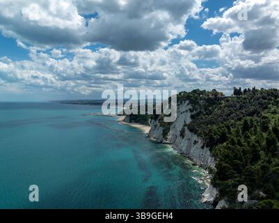 Vista aerea della splendida costa di Sirolo, Italia. Scogliere spettacolari sorgono dal mare turchese, con vegetazione lussureggiante sotto un cielo parzialmente nuvoloso Foto Stock