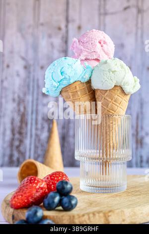Tre coni da gelato collocati in un bicchierino decorativo in vetro. In primo piano, fragole e mirtilli fuori fuoco, in secondo piano, due addit Foto Stock
