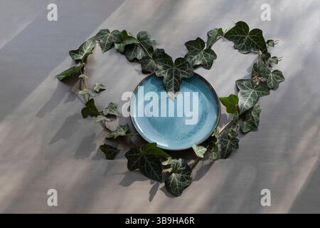 Una forma a cuore fatta di foglie di edera su sfondo grigio e un piatto blu al centro, una decorazione simbolica sostenibile sul tema dell'amore Foto Stock