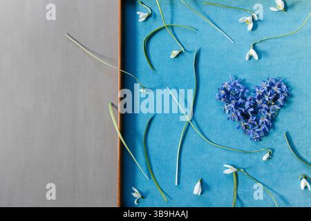 Un cuore fiorito di blu e viola fatto di stelle blu giace in una cornice di legno circondata da gocce di neve bianche in un'elegante disposizione floreale sul tema Foto Stock