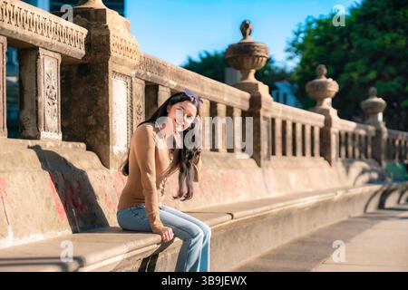 Giovane donna latina con capelli lunghi e outfit casual sorridente mentre siede su una panchina di pietra su Reforma avenue a città del Messico. Foto Stock